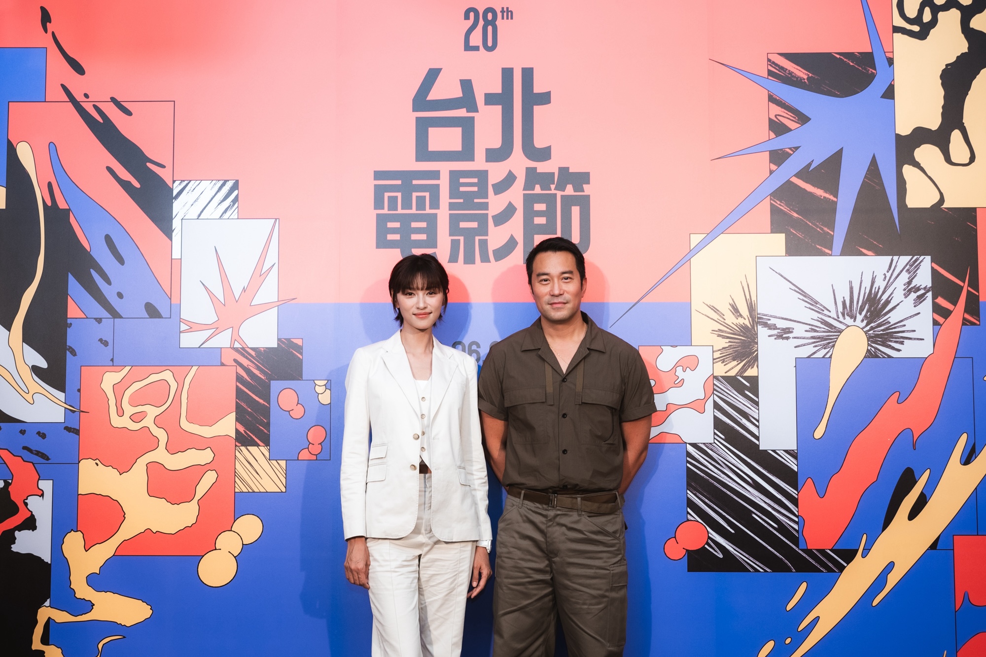 一名身穿白色西裝的女子與一名身穿深色襯衫的男子，並肩站在第28屆台北電影節彩色主視覺牆前合影